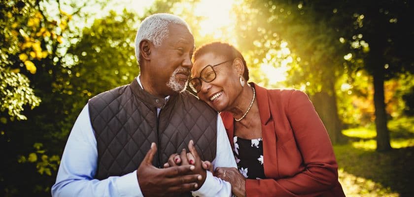 An elderly couple shares a joyful moment outdoors, smiling and holding hands in a sunlit park.