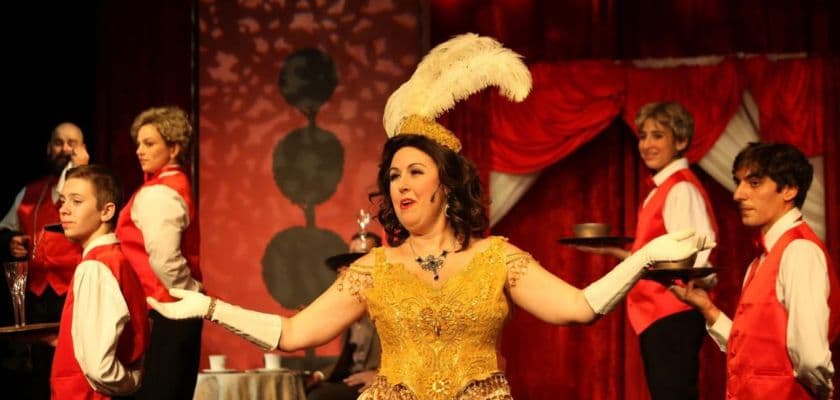 A performer in a glamorous yellow dress and feathered headpiece gestures dramatically, surrounded by cast members in a theatrical setting.