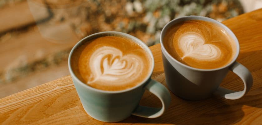 Two cups of latte with heart-shaped foam art sit on a wooden surface.
