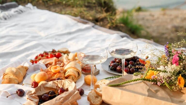 A picnic spread with wine glasses, fruits, pastries, and a bouquet of flowers on a white blanket by the water.
