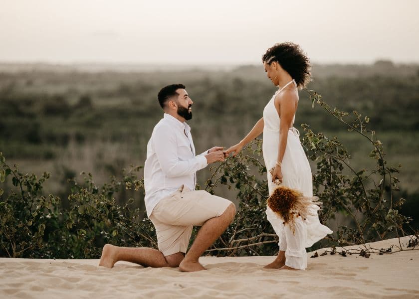 A man kneels in the sand, holding hands with a woman in a flowing white dress, with a scenic landscape in the background. A man kneels in the sand, holding hands with a woman in a flowing white dress, with a scenic landscape in the background.