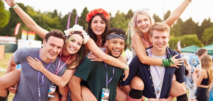 A group of six friends smiles and poses for a photo at a festival, with vibrant accessories and a lively outdoor setting.
