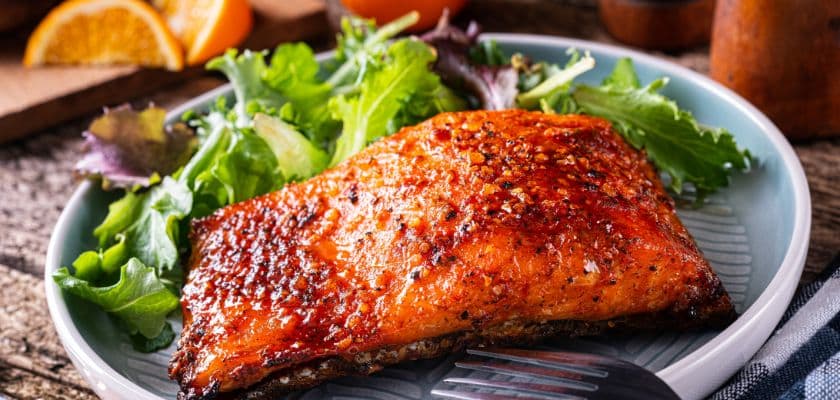 A glazed salmon fillet sits on a plate with fresh greens and orange slices.