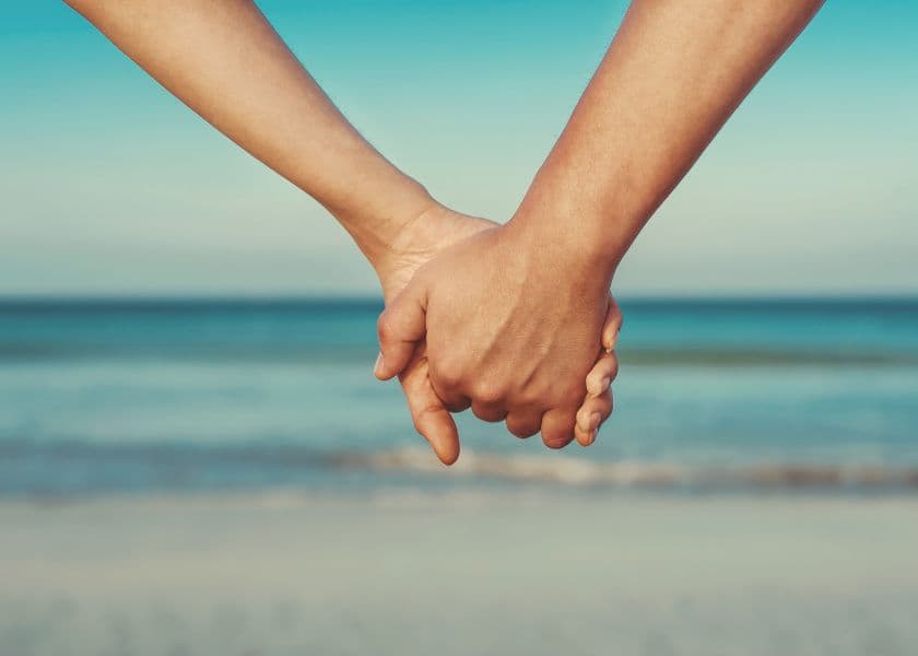 Couple holding hands on the beach with the ocean in the background.