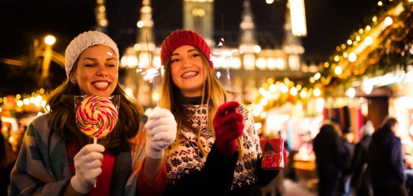 two friends in winter hats smiling and holding sparklers and a lollipop at festive outdoor holiday market
