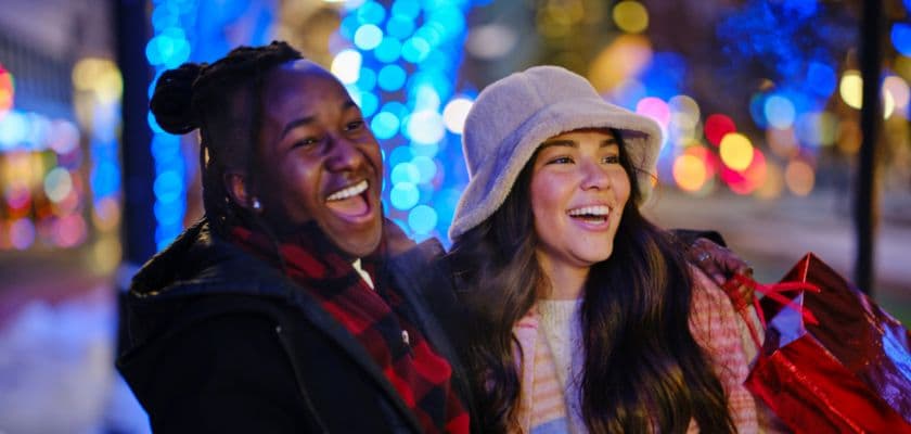 two friends smiling and laughing while shopping at night surrounded by colorful holiday lights
