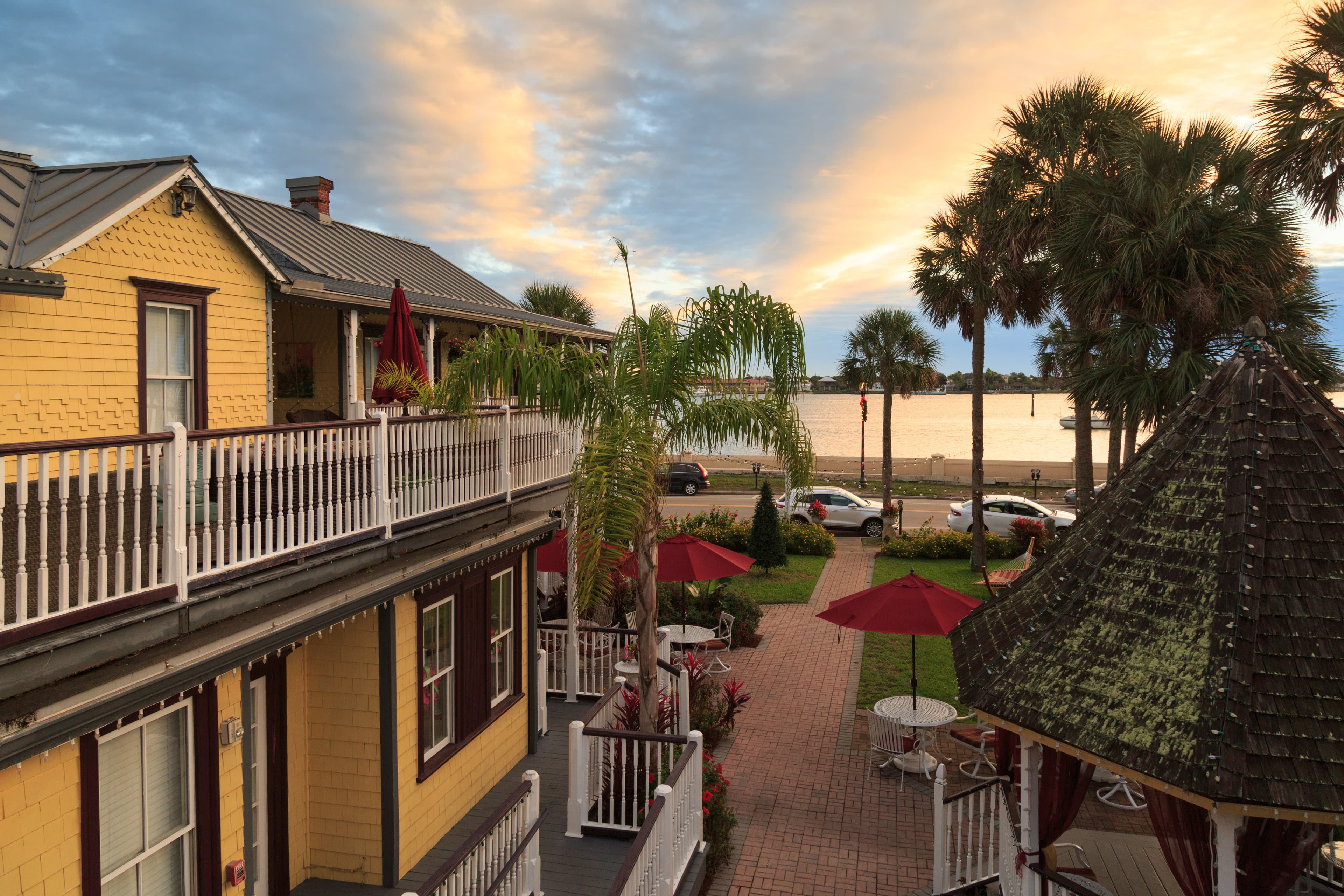 A historic yellow building with white railings overlooks a waterfront with palm trees and a gazebo at sunset.