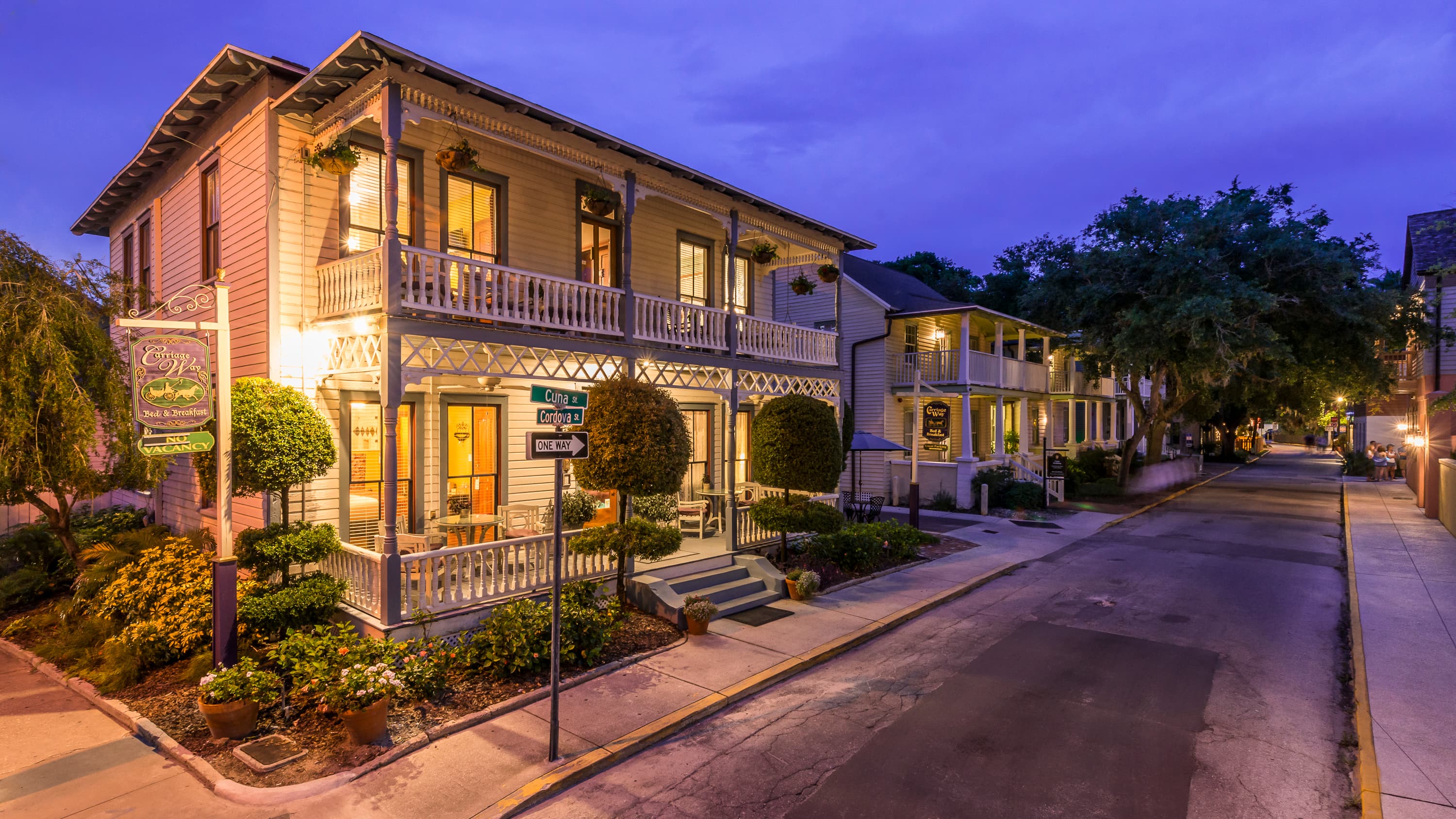 A large, historic building with balconies and illuminated windows stands at dusk, flanked by a street sign and lush greenery.