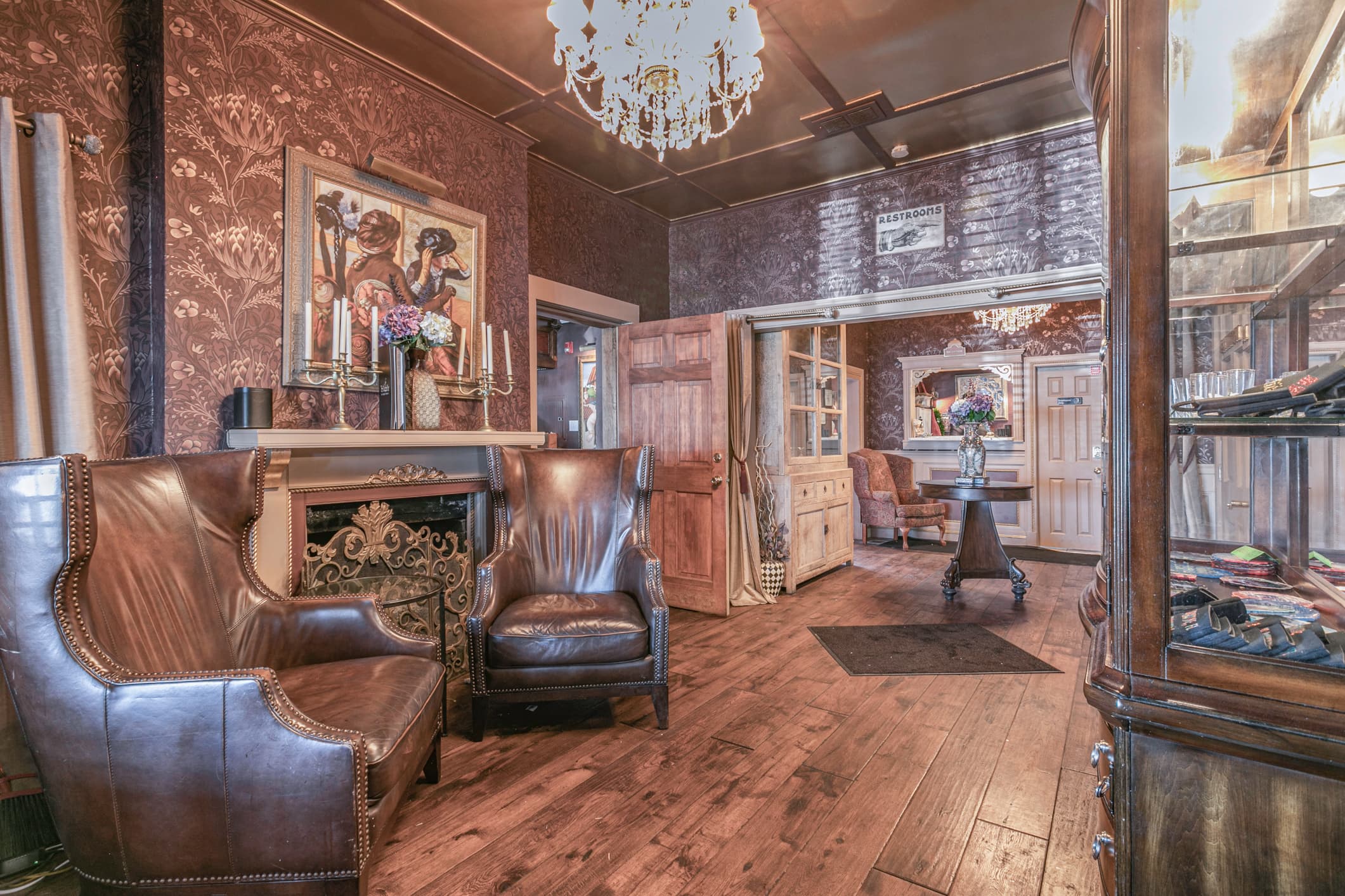 An ornately decorated room features two leather armchairs by a fireplace, a chandelier, and dark wallpaper with intricate patterns.
