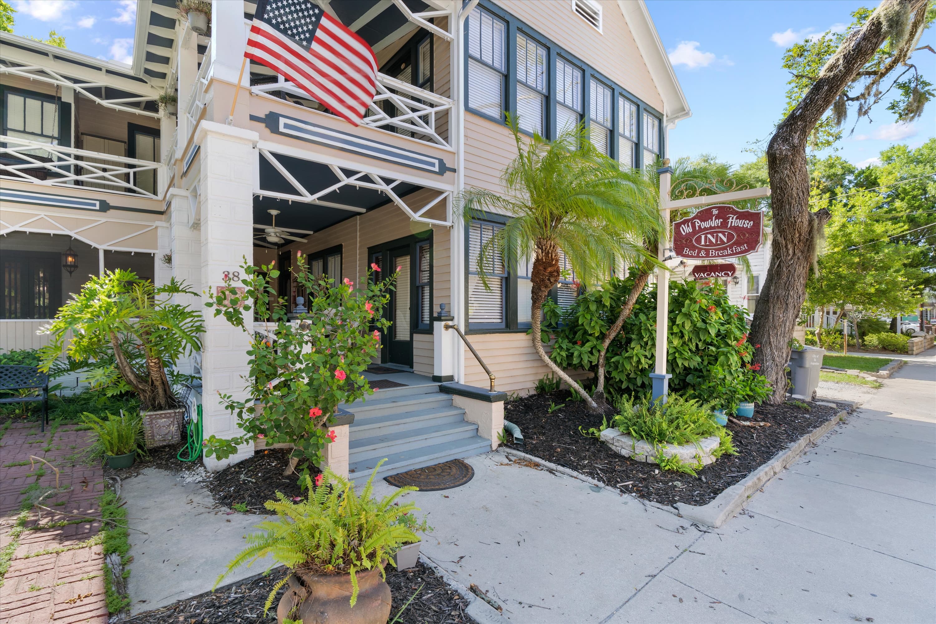 A cream-colored historic inn with an American flag displayed on its balcony stands beside a sidewalk and lush tropical landscaping under a bright sky.