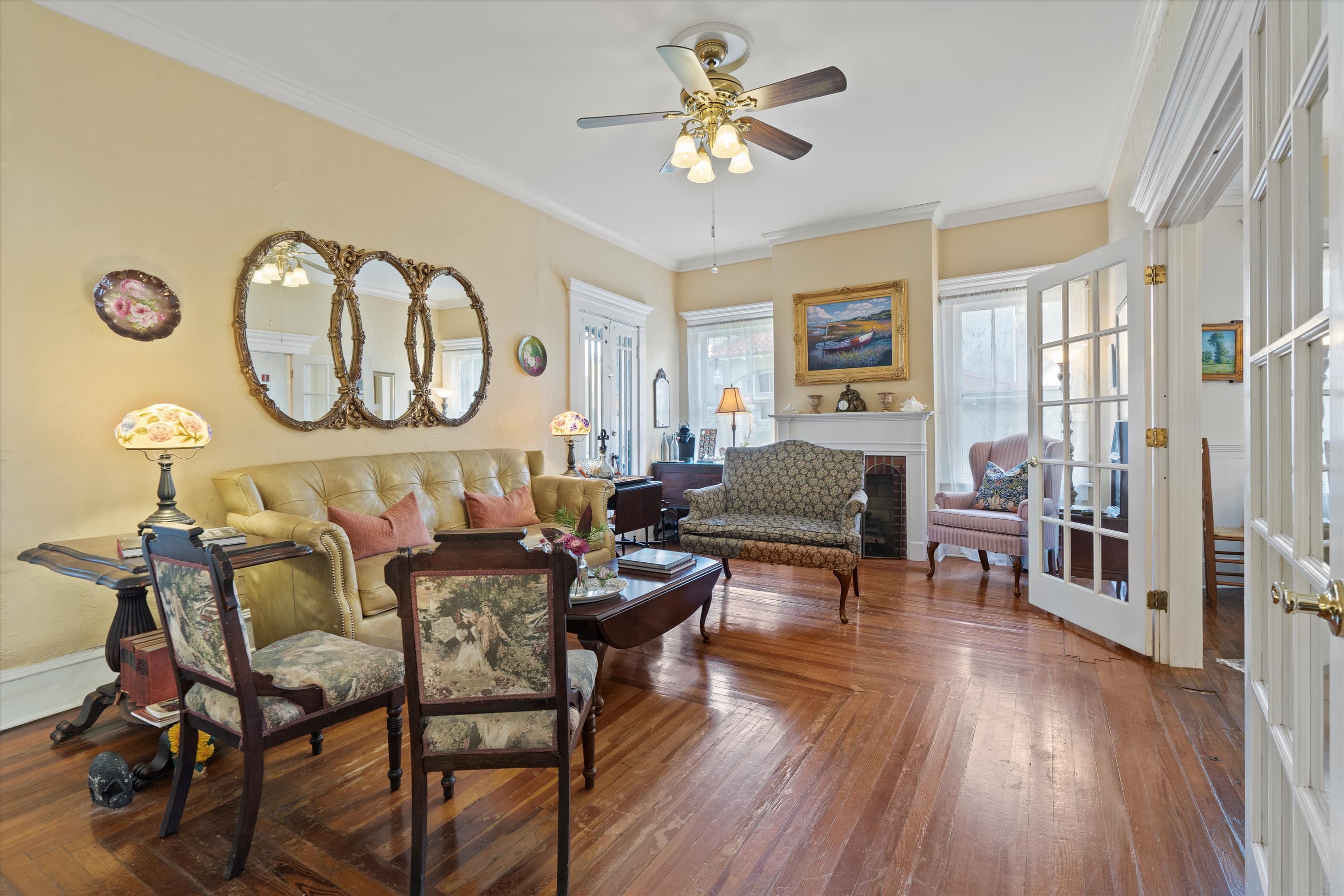 A bright, elegant living room features polished hardwood floors, a yellow sofa, an ornate coffee table, and multiple mirrors on the wall.