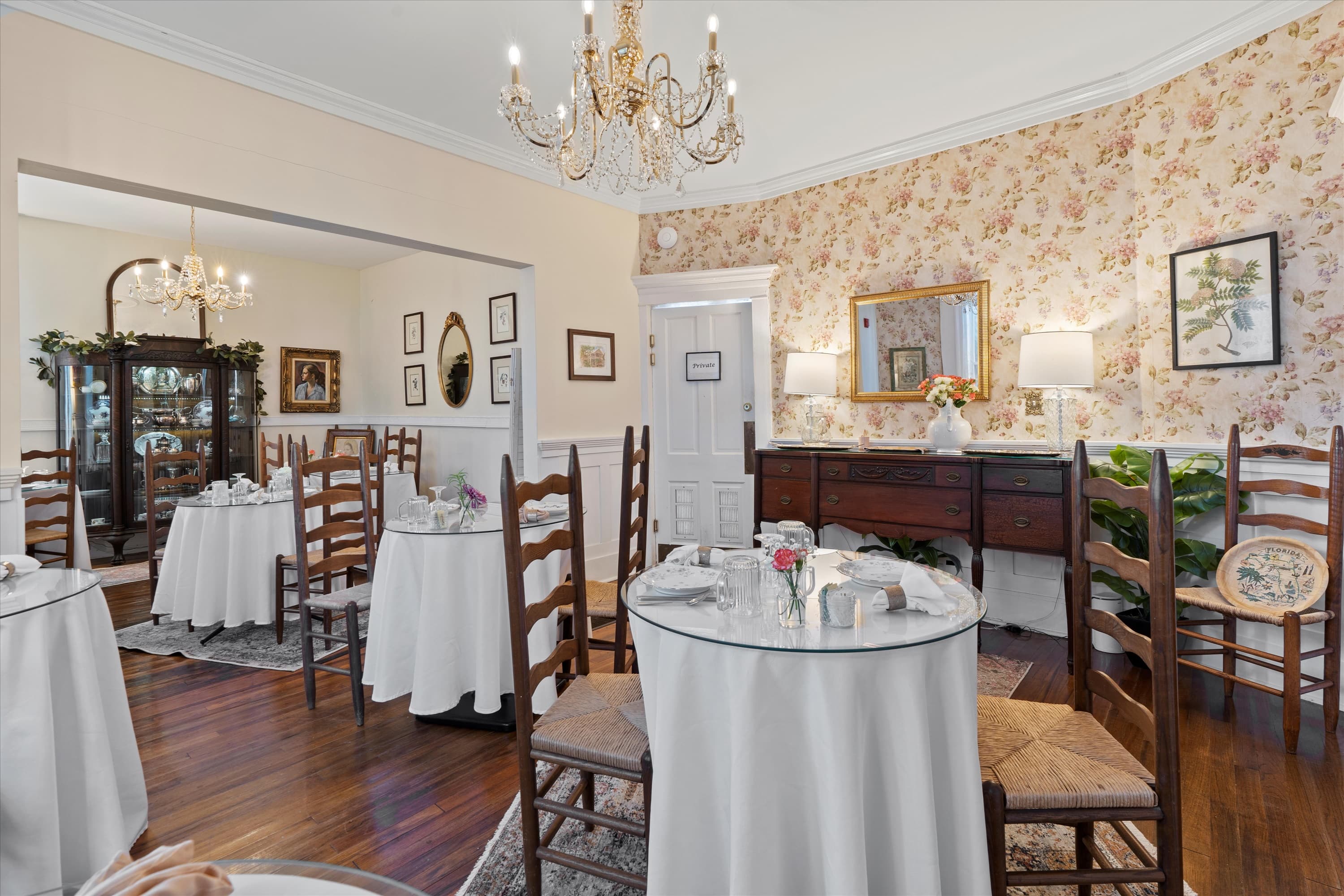 An elegant dining room features multiple round tables with white tablecloths, a chandelier, and floral wallpaper on one wall.