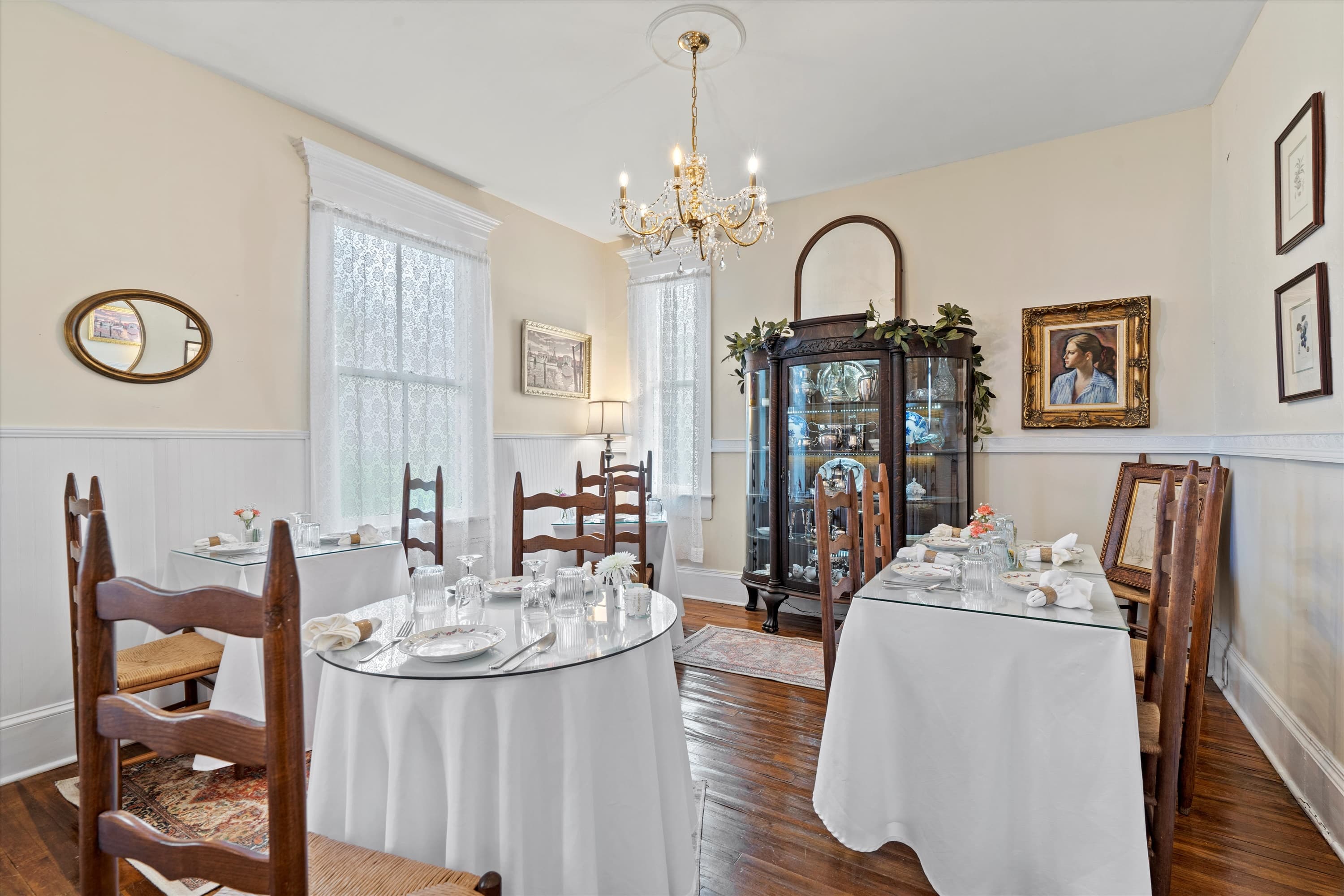 An elegant dining room features several white-clothed tables, dark wooden chairs, a glass curio cabinet, and a chandelier, all set against light-colored walls.