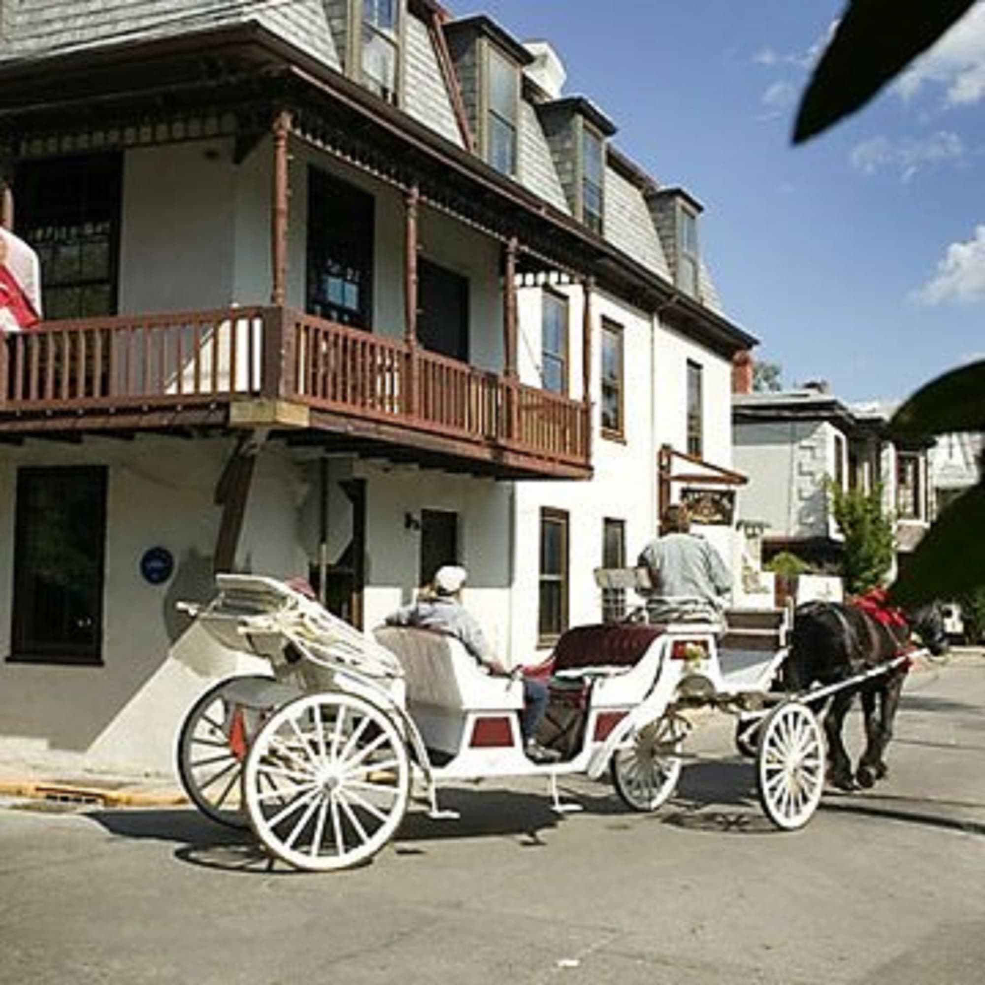 A white horse-drawn carriage with two people on board moves past a multi-story white building with balconies.
