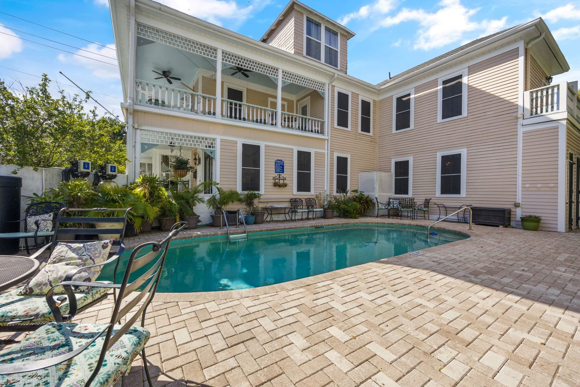 A sunny outdoor patio features a blue swimming pool surrounded by brick paving, lounge chairs, and a light-colored multi-story building with balconies.
