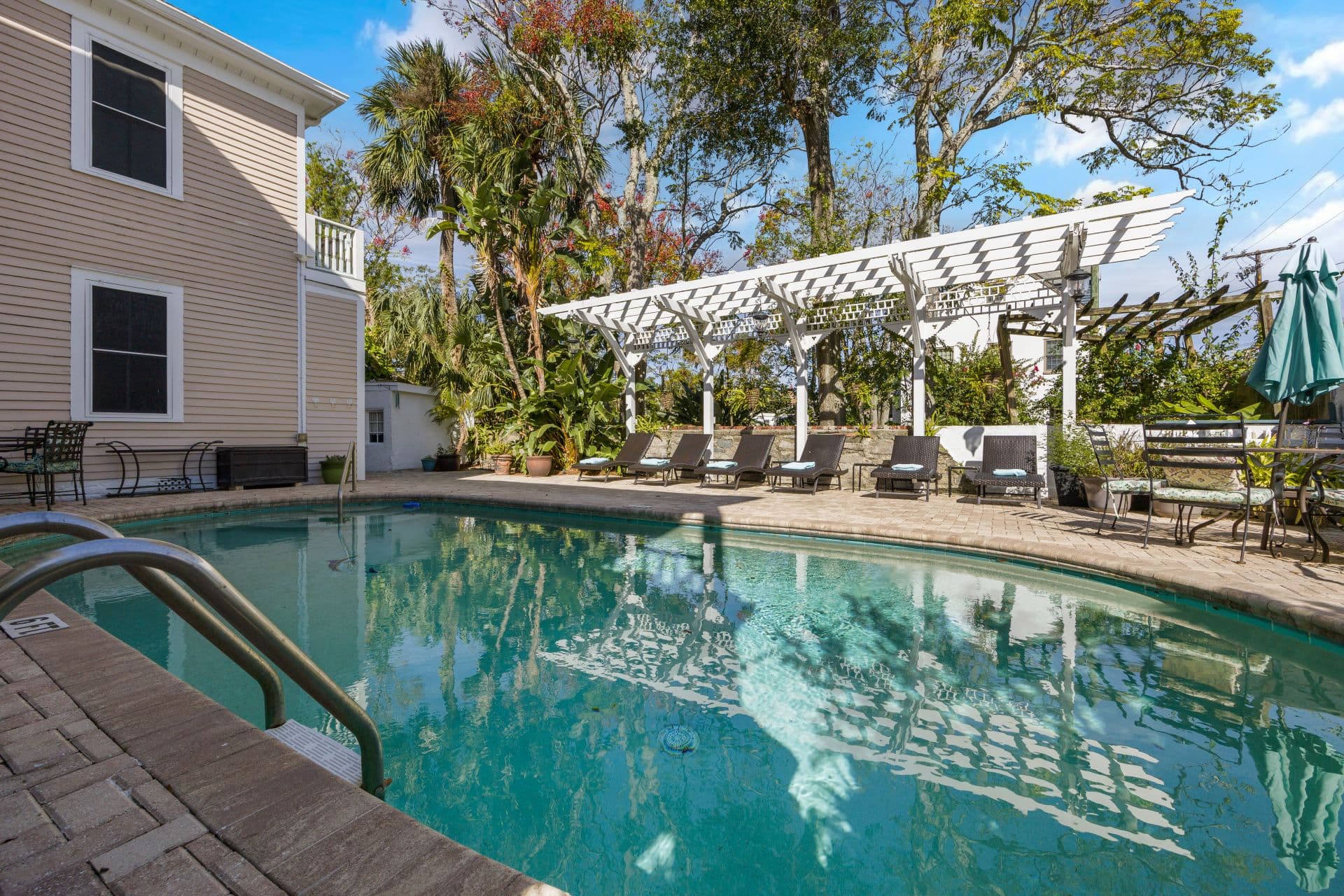 A refreshing swimming pool with a metal ladder is surrounded by a brick patio, lounge chairs under a white pergola, and lush greenery.
