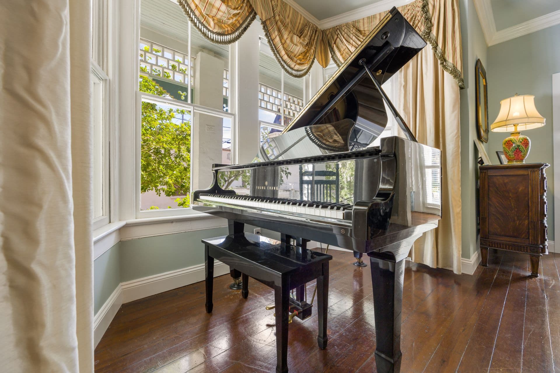 A shiny black grand piano sits on a polished wooden floor in a room with large windows and ornate drapes.
