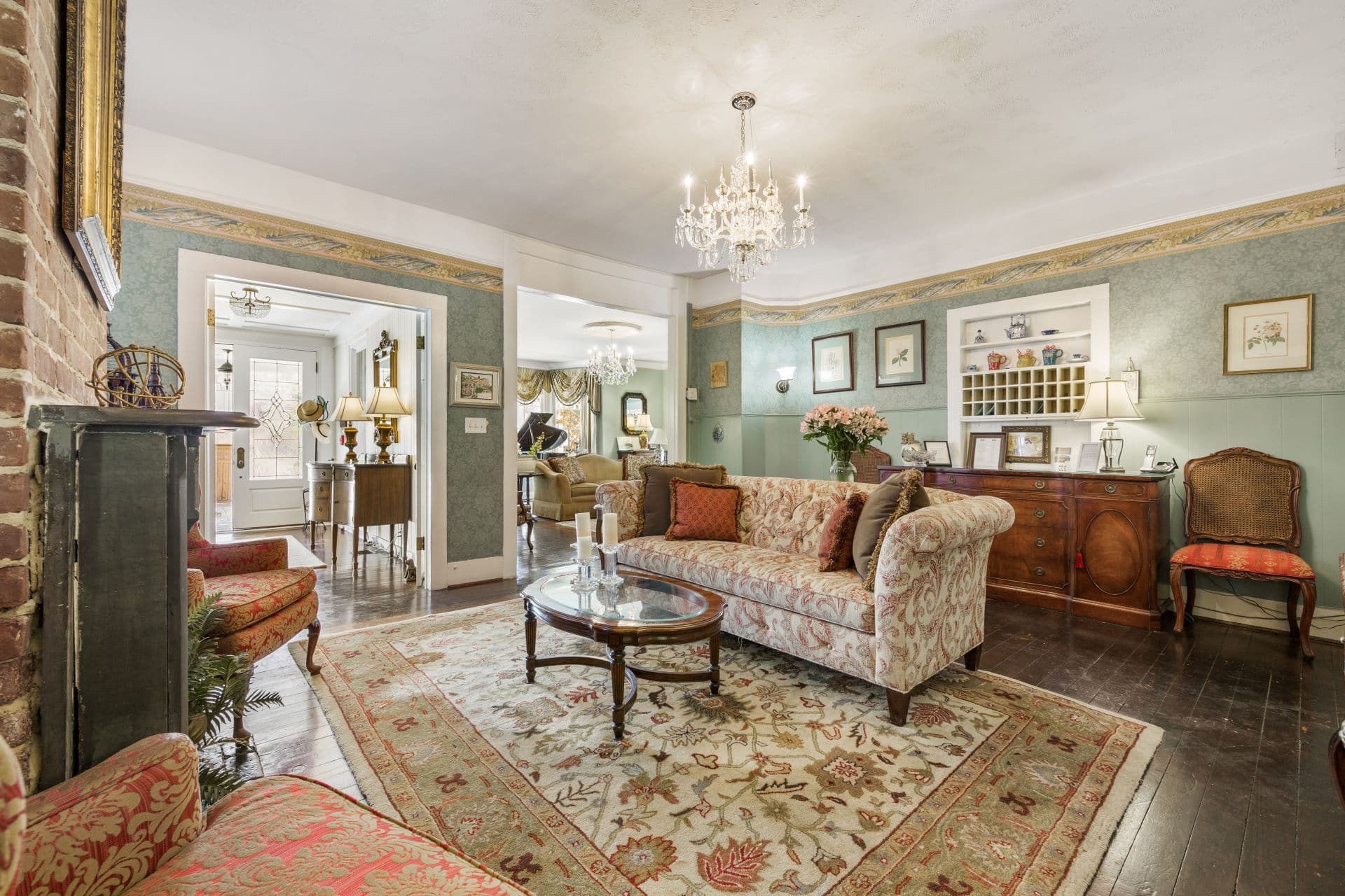 A formal living room with a patterned sofa, a crystal chandelier, and an ornate rug, with a glimpse into another room with a grand piano
