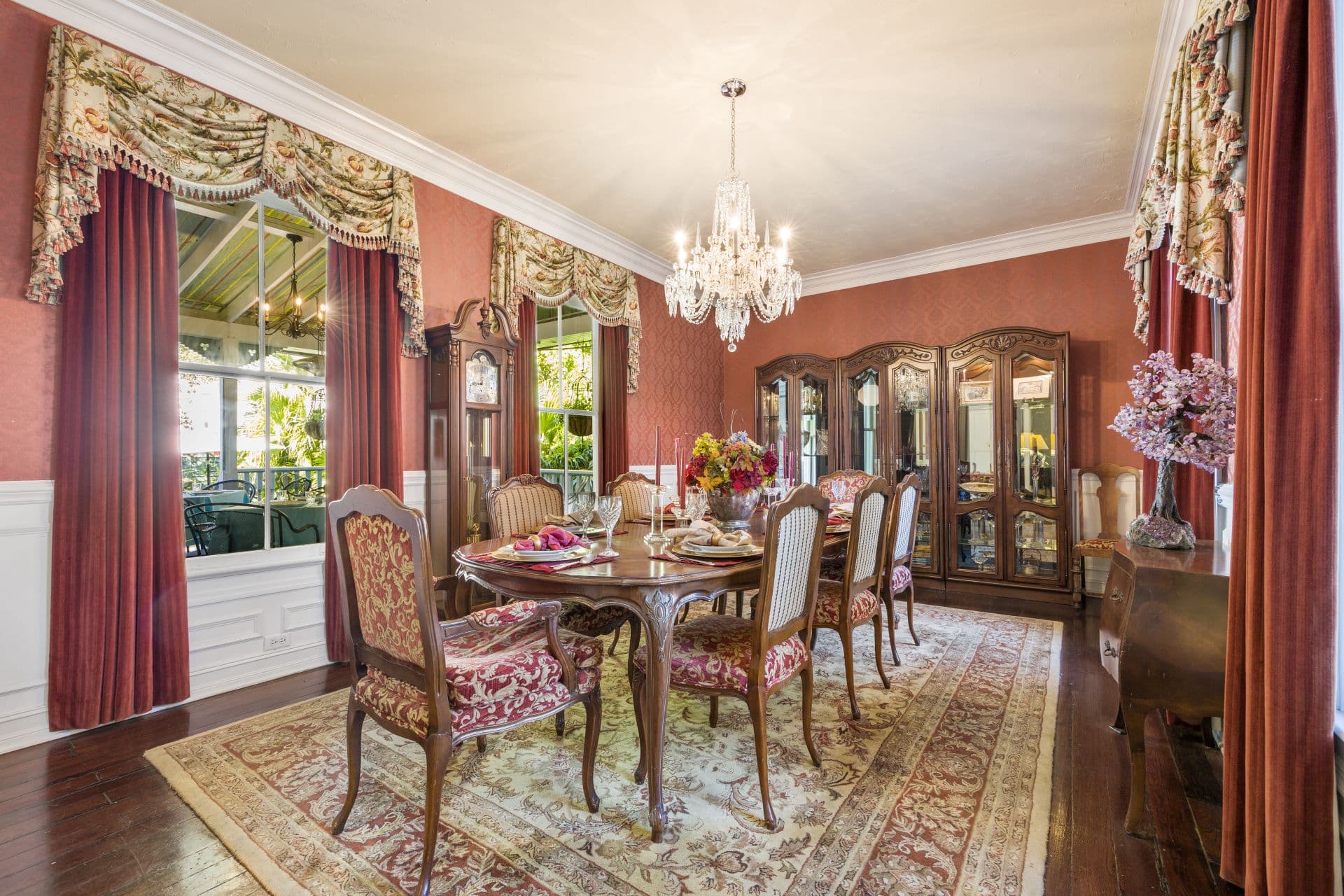 An ornate dining room with red walls, velvet curtains, a crystal chandelier, and a large wooden dining table set for dinner.