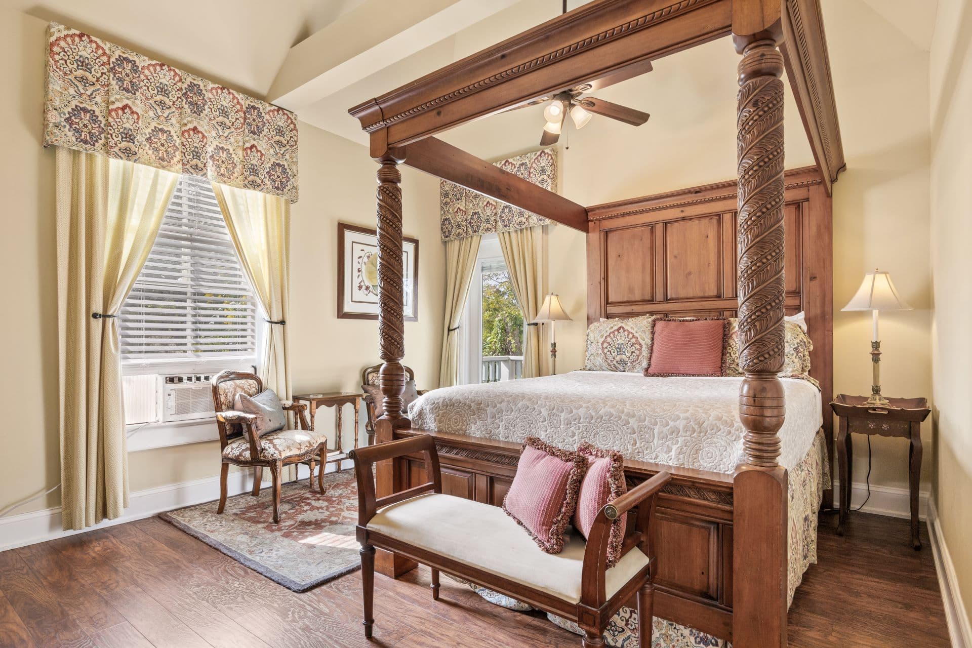 A luxurious bedroom features a grand wooden four-poster bed with patterned bedding, two windows with drapes, and a seating area.