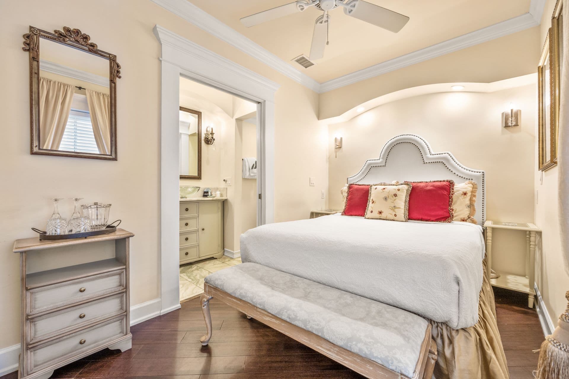 A well-lit bedroom features a large white bed with a tufted headboard and red pillows, a ceiling fan, and an open doorway leading to a bathroom.