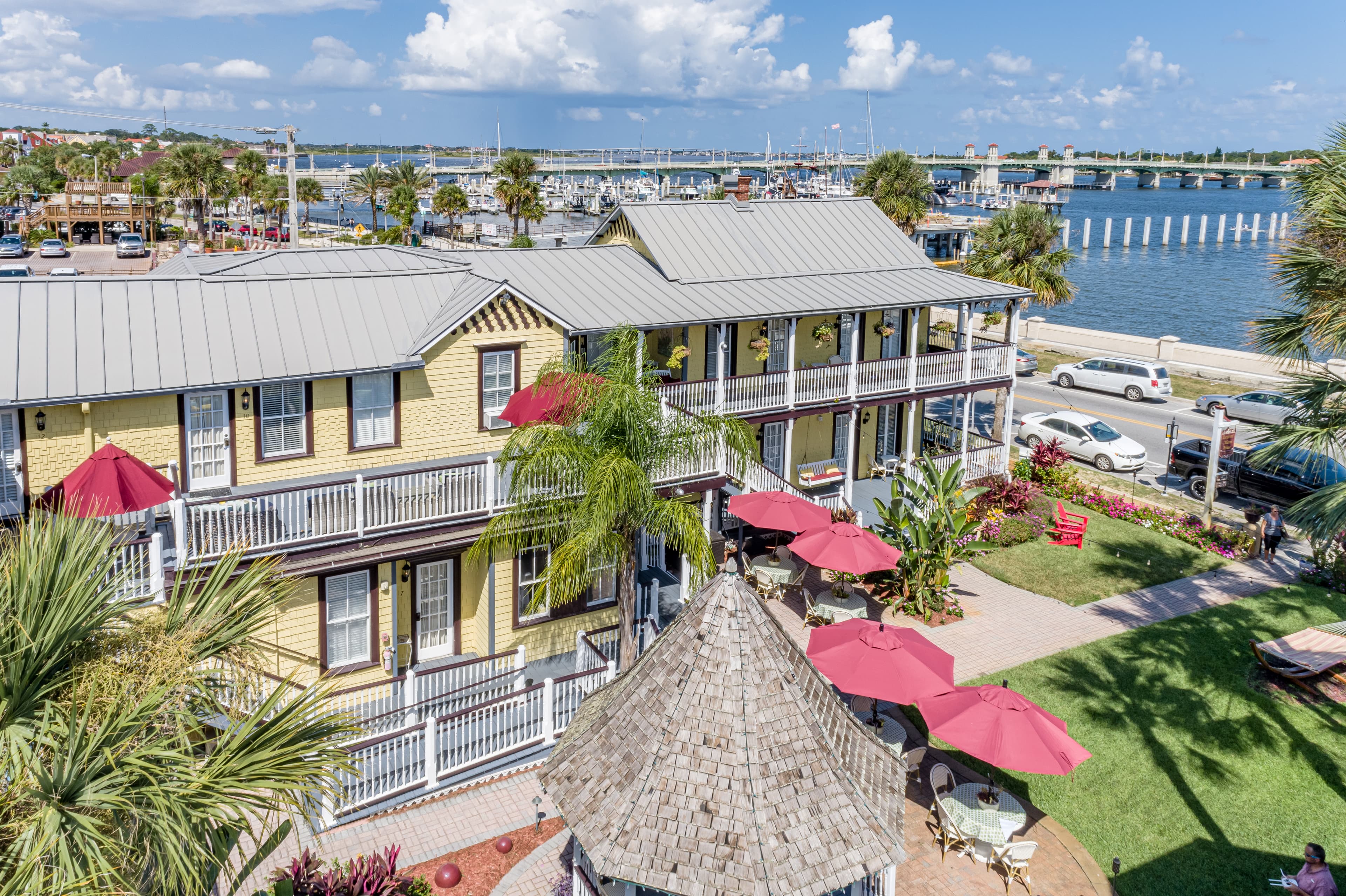 An aerial view of a yellow building complex with gray roofs, red umbrellas, and a body of water with a bridge in the background.