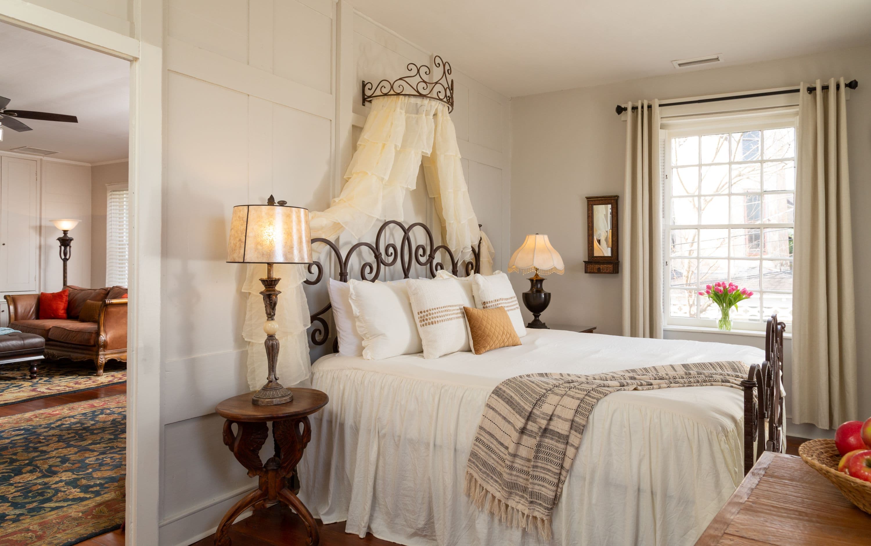A bedroom with a metal bed frame, white bedding, a canopy, and a window, with a living area visible beyond an open doorway.