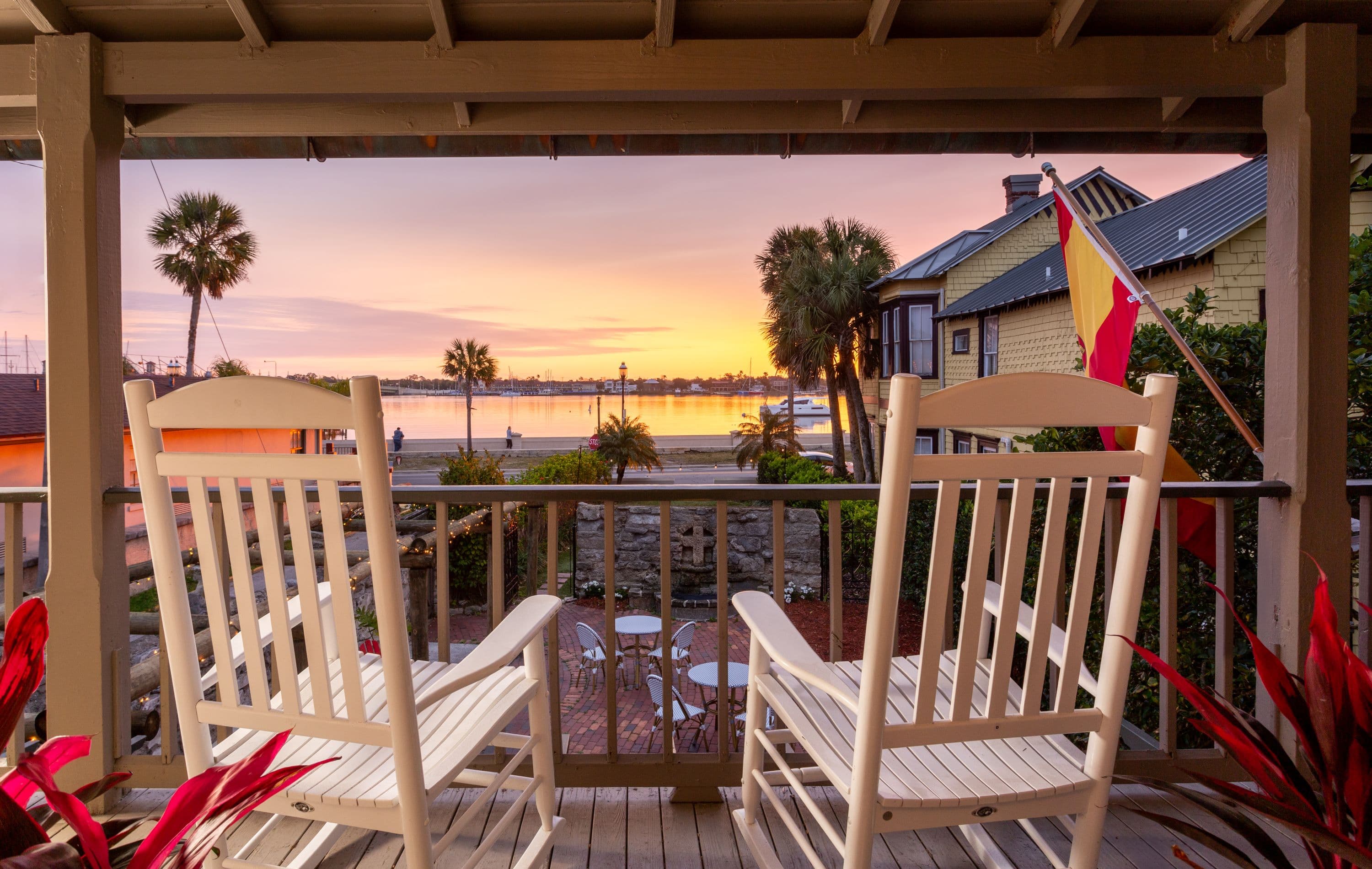 Two rocking chairs on a balcony overlooking a water view at sunset, with a building and palm trees.