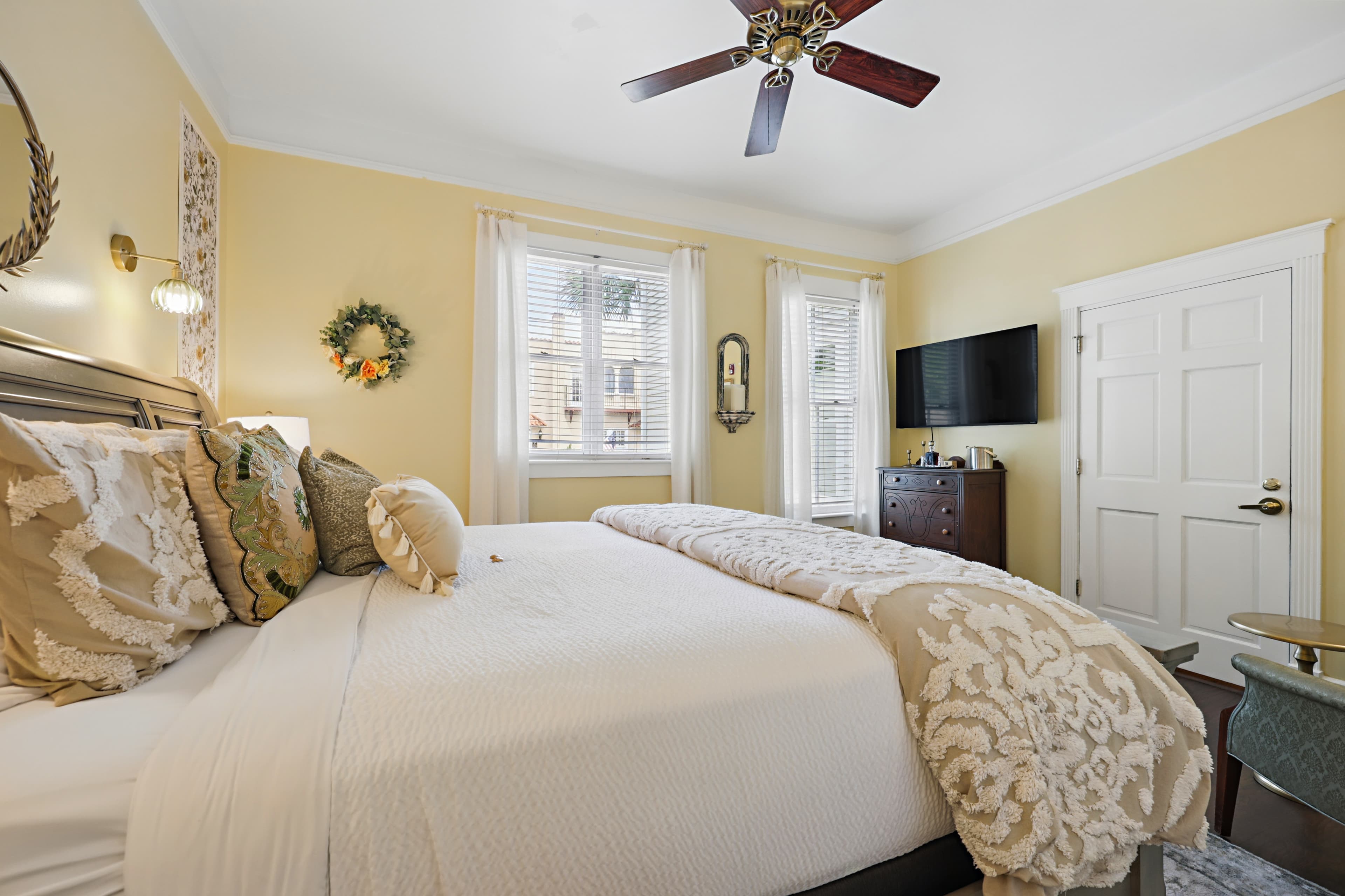 Beautiful cream colored guest room with a king bed, smart tv on the wall, double windows, and a ceiling fan.