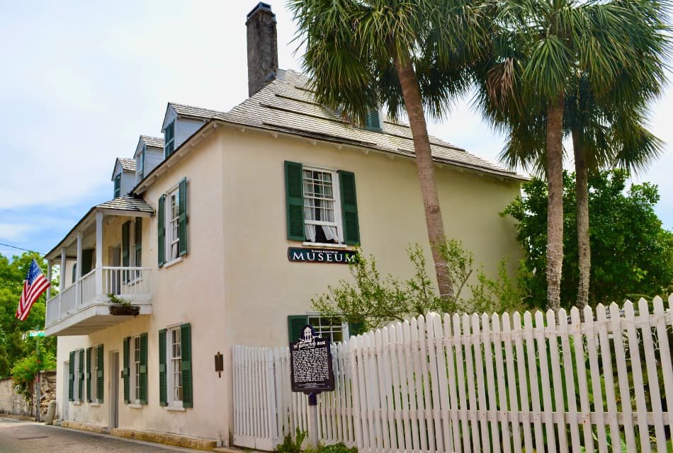 Old two-story building painted white with green shutters next to palm trees and white-picket fence