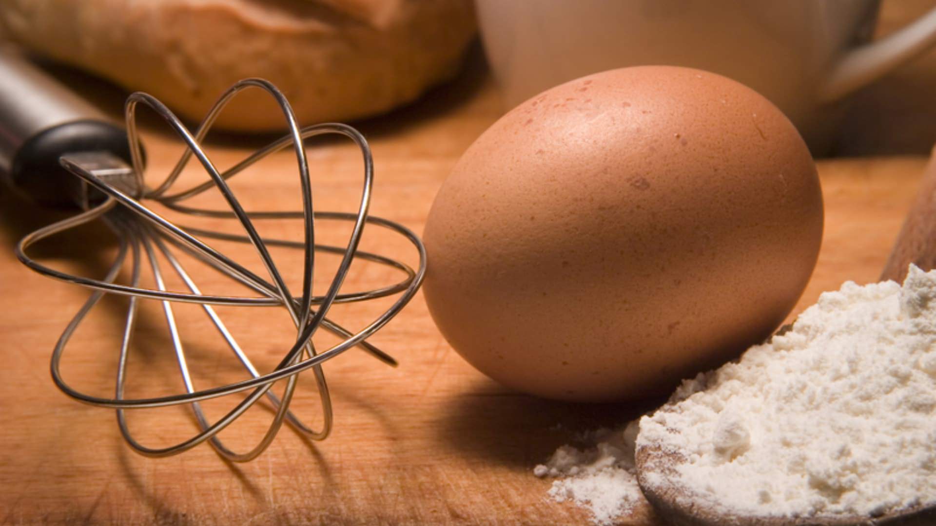 Close up view of brown egg, whisk, and flour on wooden cutting board