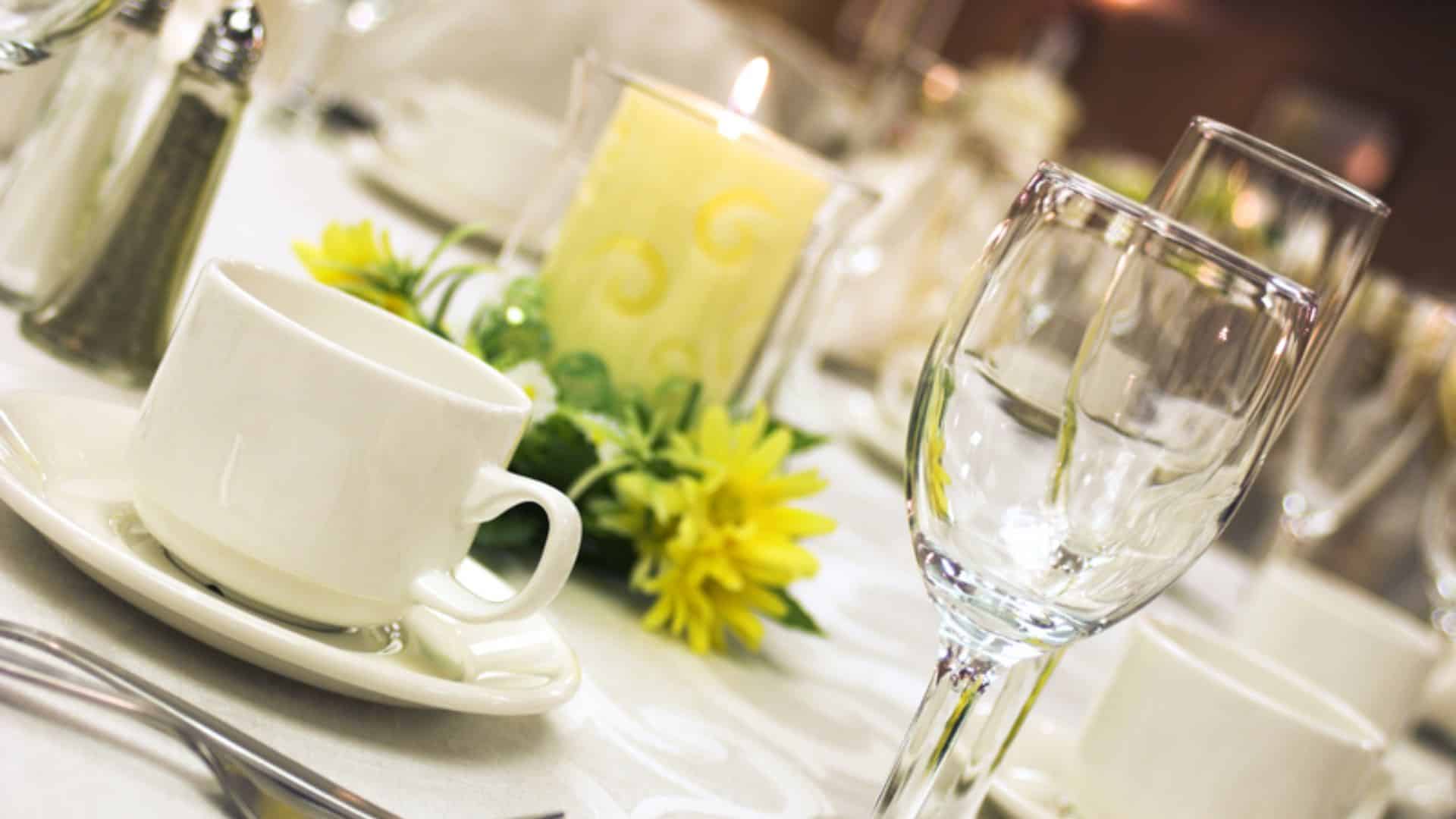Close up view of glasses, white coffee cups with saucers, and a yellow candle on a white tablecloth