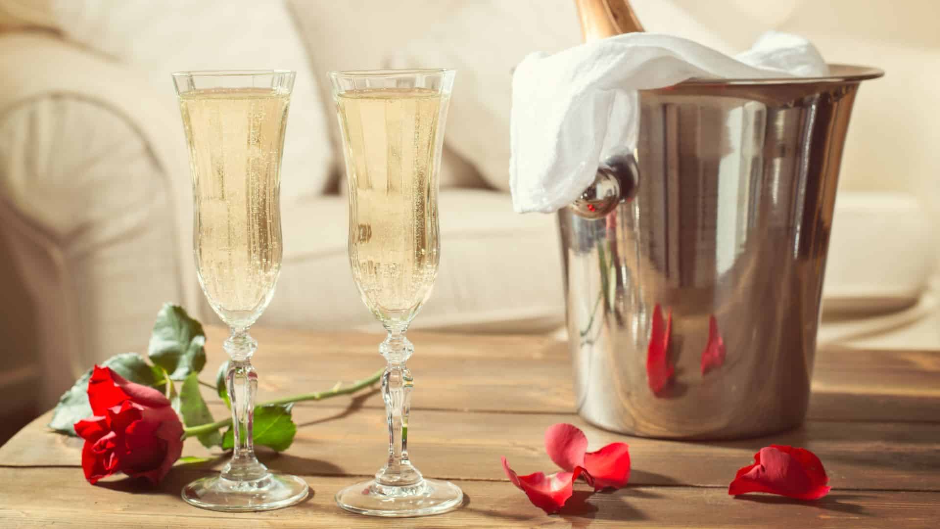 Two glasses full of Champagne next to a red rose and wine bucket on a wooden table