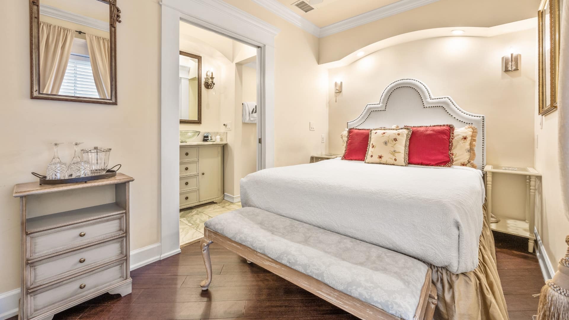 Bedroom with cream walls, hardwood flooring, white upholstered headboard, white bedding, and view into bathroom