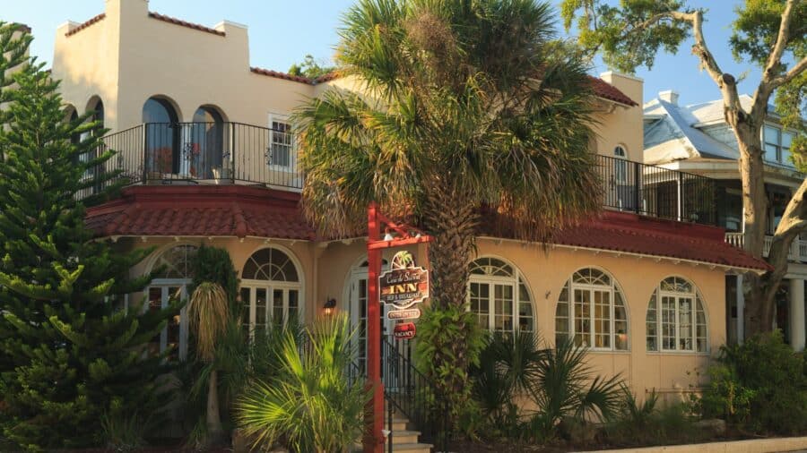 Exterior view of property painted light tan with white trim, porch on lower and upper levels, green vegetation, and palm trees