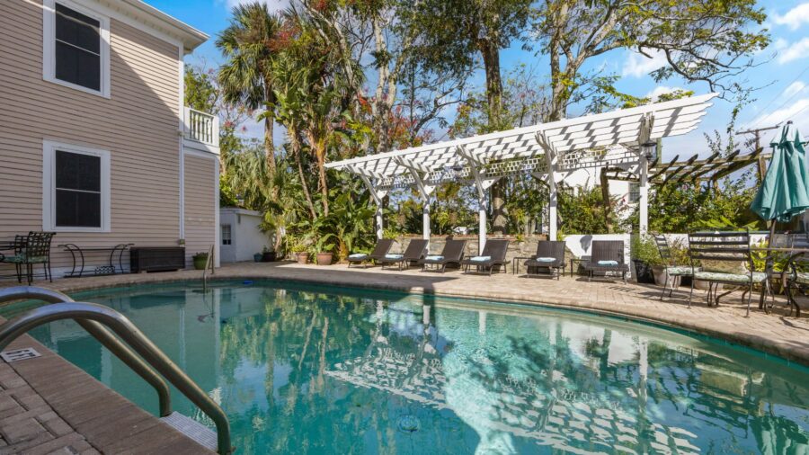 Exterior view of property painted cream with white trim, pool, paver pool deck with lounge chairs, white pergola, and large trees