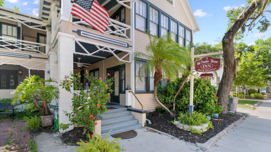 Exterior view of property painted cream with blue and green trim, porch on lower and upper levels, paver patio, bushes, and palm trees