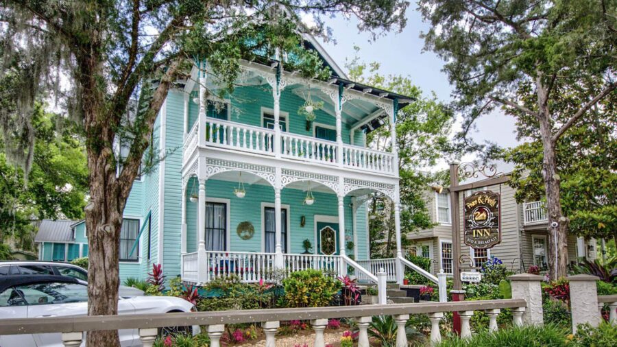 Exterior view of property painted light turquoise with white trim, porch on lower and upper levels, bushes, and palm trees