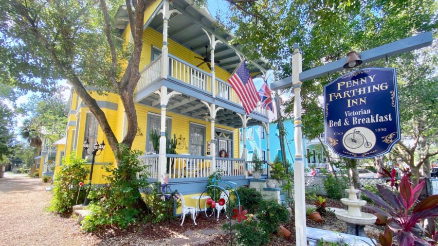 Exterior view of property painted yellow with blue trim, porch on lower and upper levels, water fountain, green bushes, and large trees