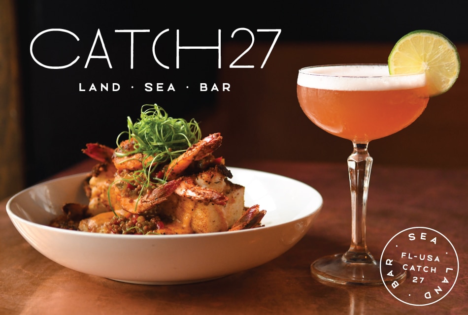 White bowl with fancy seafood dish next to a cocktail drink with a slice of lime on a wooden table and the words Catch 27, which is the name of the restaurant
