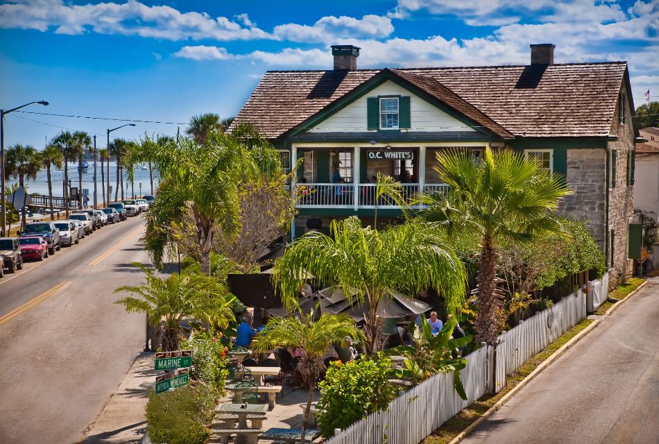 Exterior view of a restaurant in a two-story stone building and patio with green vegetation and palm trees