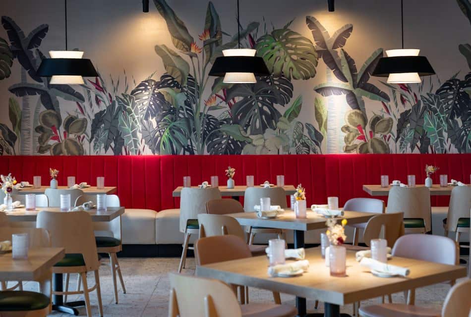 Restaurant dining area with large floral mural, tile flooring, light wooden tables, and light wooden chairs with cushions