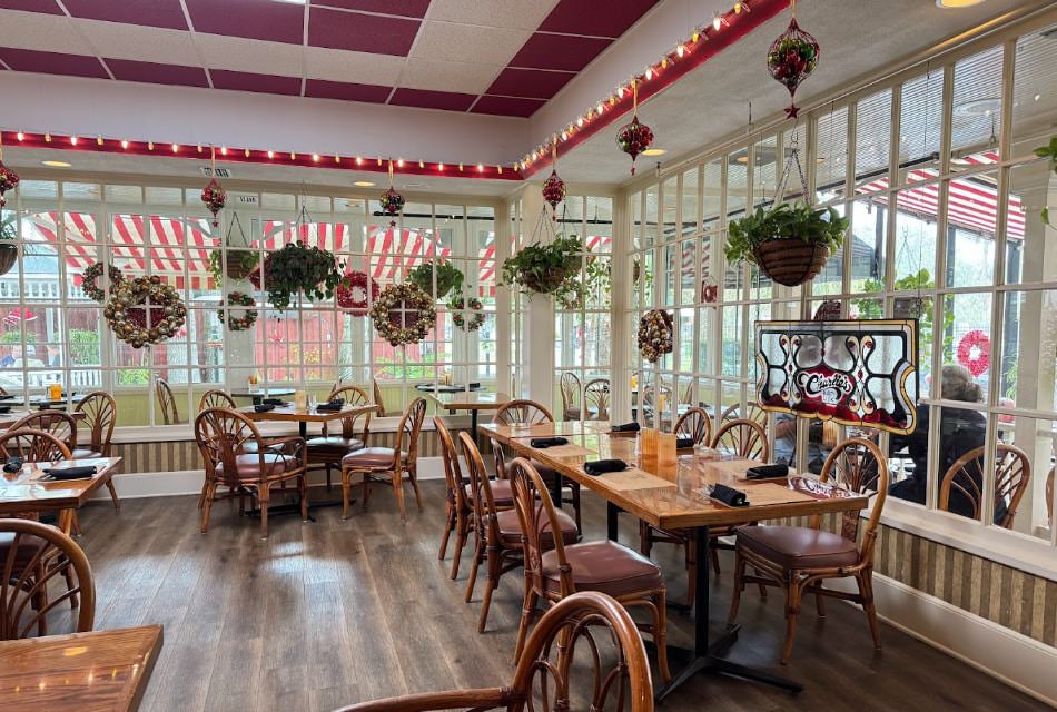 Restaurant dining area with multiple large windows, white trim, hardwood flooring, wooden tables, and wooden chairs with cushions