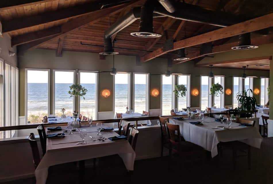 Restaurant dining area with multiple large windows, white trim, carpeting, tables with white tablecloths, wooden chairs, and view of the beach